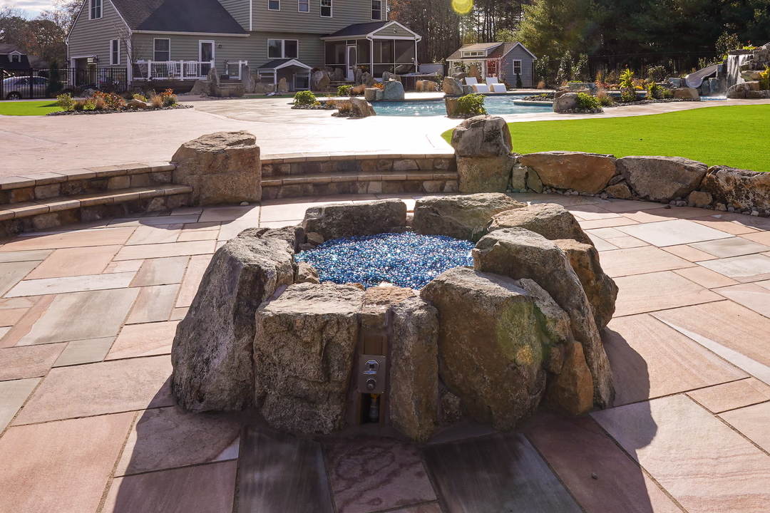 Willow R - Port Republic, NJ - This custom outdoor living project features a natural stone waterfall cascading over large boulders into a gunite pool. The waterfall includes a blue glass pebble bed at its base, creating a visually striking contrast against the rugged stone. The surrounding area is paved with large, light-colored stone tiles, and the design integrates seamlessly with the landscaped backyard, which includes a well-maintained lawn, additional rock formations, and a small pool area with a waterfall and slide.